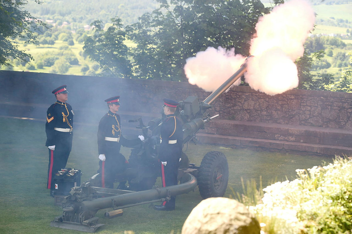 Three soldiers of the Royal Horse Artillery stand next to a large field gun as it fires. They wear ceremonial uniforms. 
